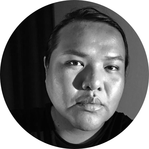A close-cropped black and white photographic headshot of a person looking directly into the camera with a neutral expression, their long black hair pulled back and lighting creating highlights and shadows on their face.