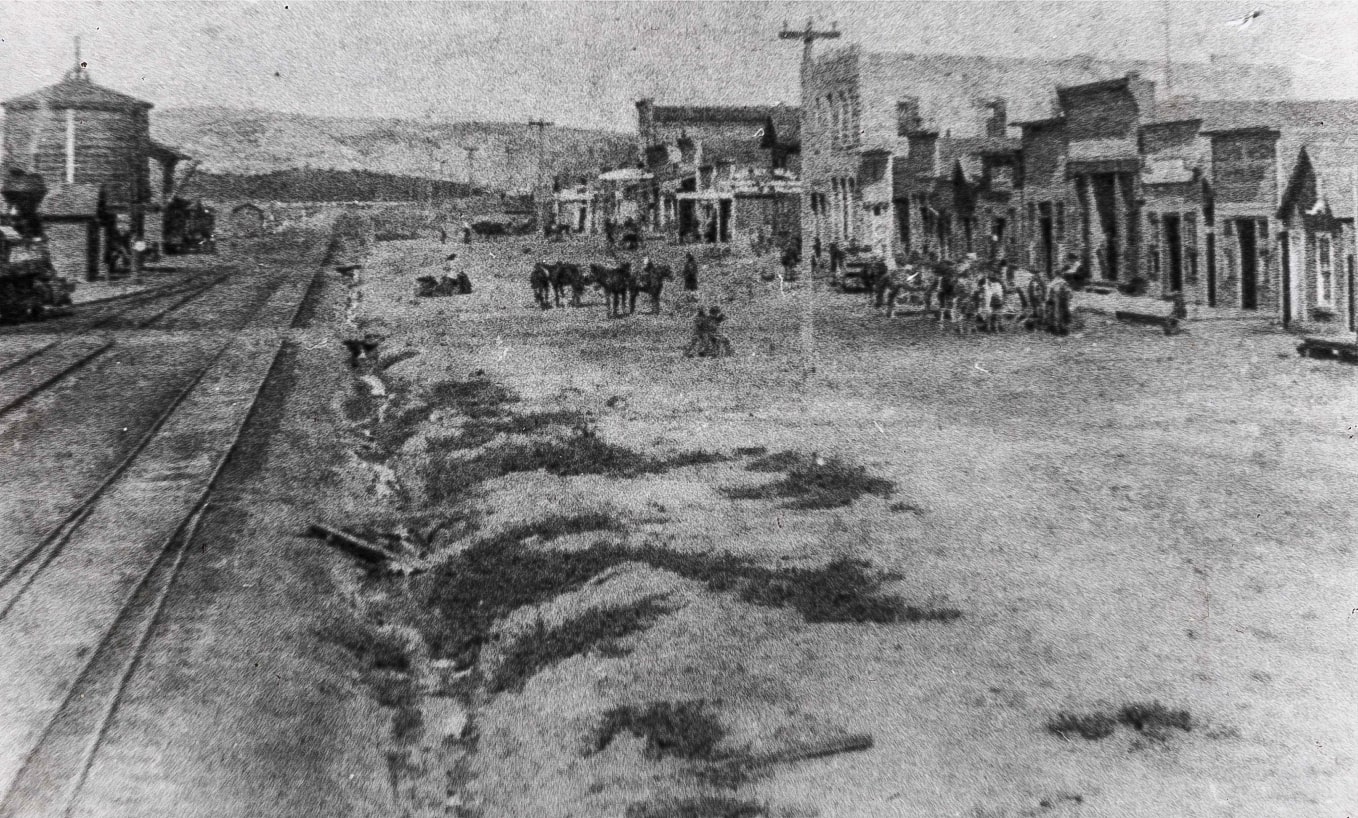 A historical black and white photograph looking down a dusty street in a small town. A row of wooden storefronts is on the right, horses gathered out front. Train tracks run parallel to the the street on the left, bordered by a small ditch. A water tower and the chimney of a steam locomotive are visible on the right edge of the image. Hills are in the distance.