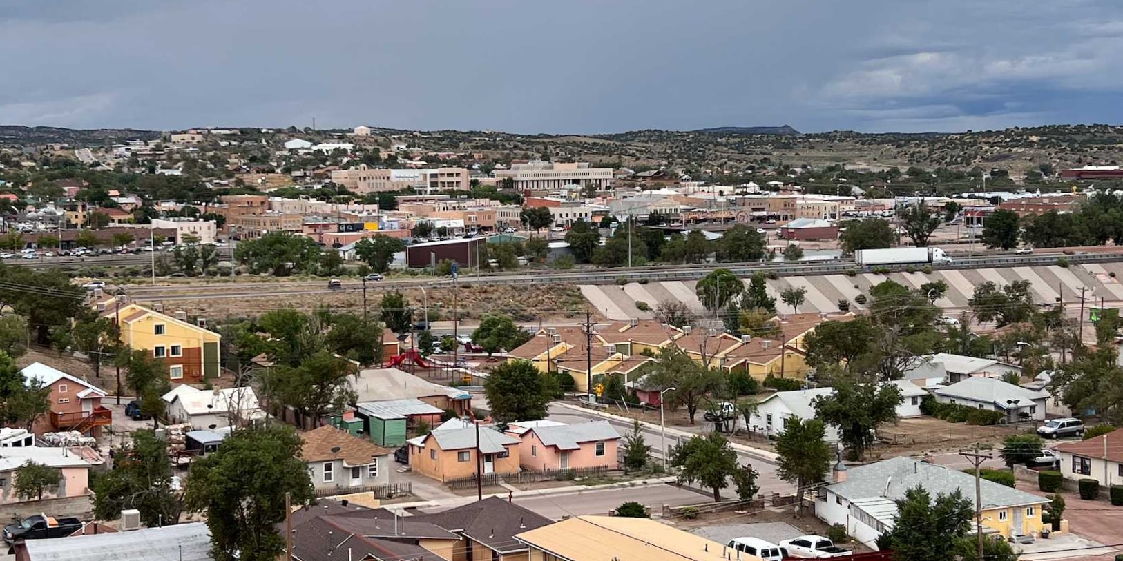 A panoramic photograph of of a small town taken from a high vantage point. In the foreground are small, colorful houses, some of which are identical and appear to be part of a development, interspersed with green trees. A raised interstate bisects the middle of the image. A dense cluster of flat-roofed, tan buildings appears behind the interstate, and building extend up the hill in the background.