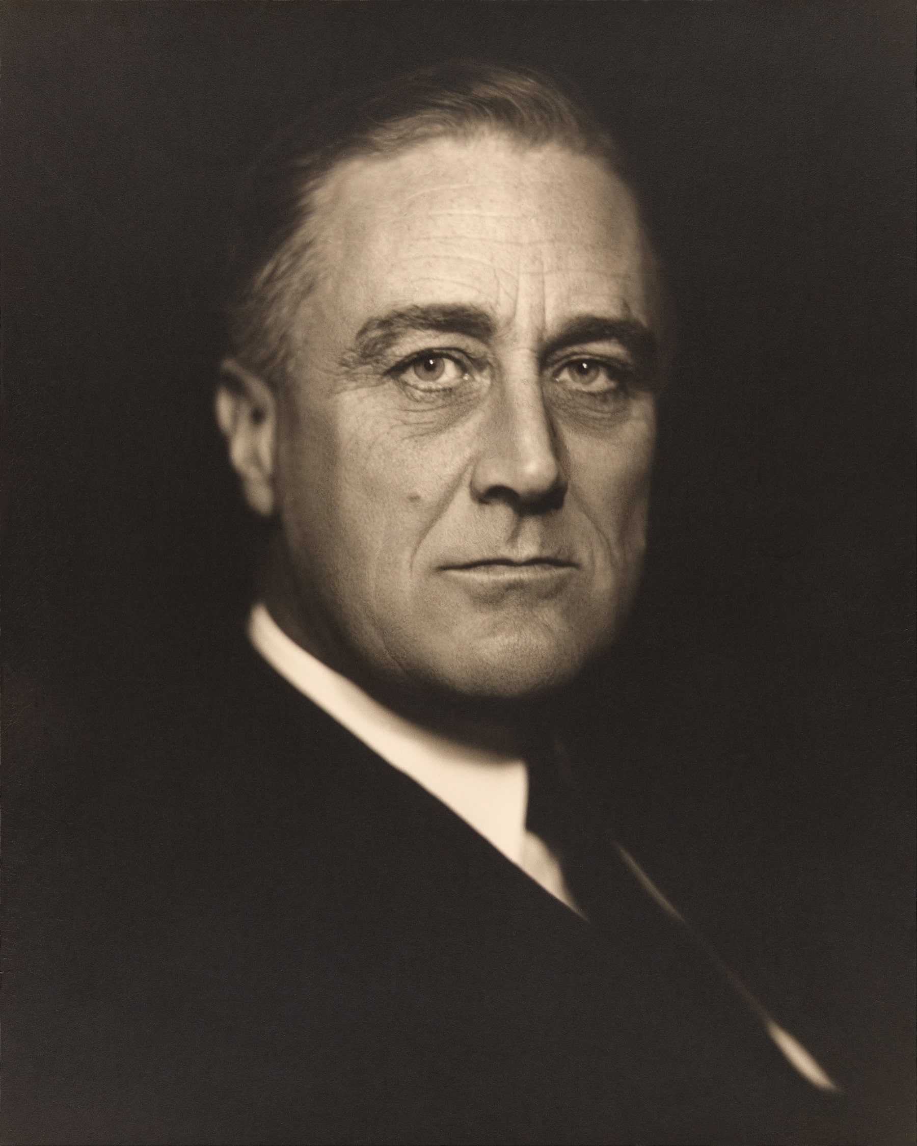 A historical black and white portrait of a middle-aged person with a serious expression, short hair, and wearing a suit and tie. The lighting is dramatic, highlighting the face against a dark background.
