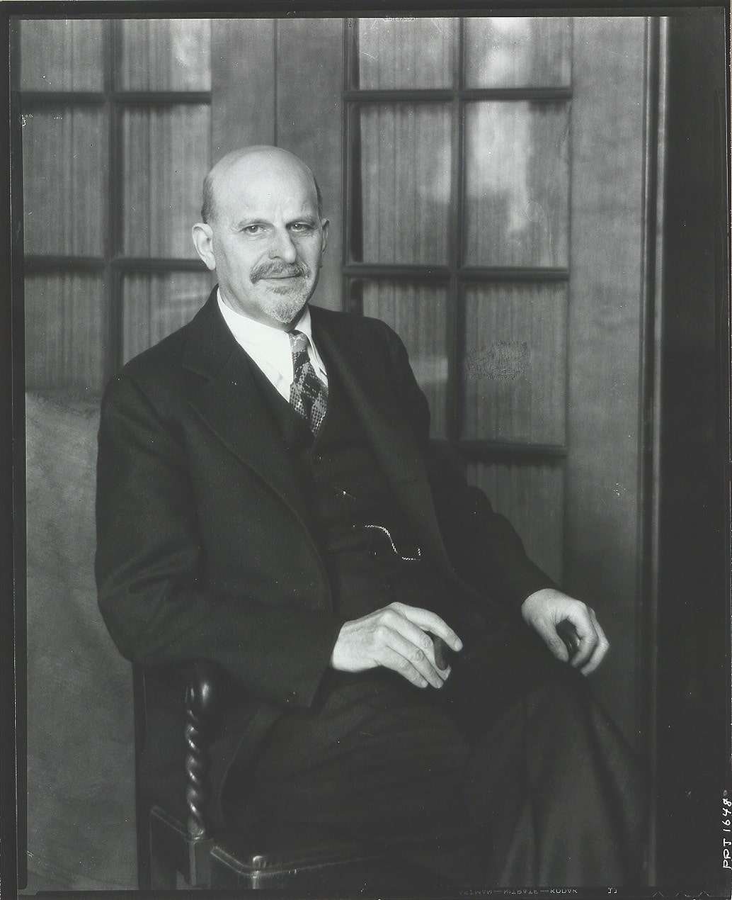 A black and white photograph of a bald man with a mustache and goatee sitting in a chair, dressed in a three-piece suit and tie. He rests one hand on his lap, holding a cigar, while the other grips the armrest. Glass-paned wooden doors are in the background. He looks directly at the camera with a slight close-lipped smile.
