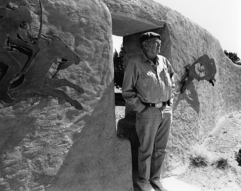 A black and white photograph of older man wearing jeans, a leather belt with a large silver buckle, a collared work shirt, a beret, and glasses, stands in the doorway of an a adobe wall. The wall features a cut sheet metal artwork on either side of the doorway, one a mounted rider and the other a bison. The man gazes into the distance, hands in his pockets.