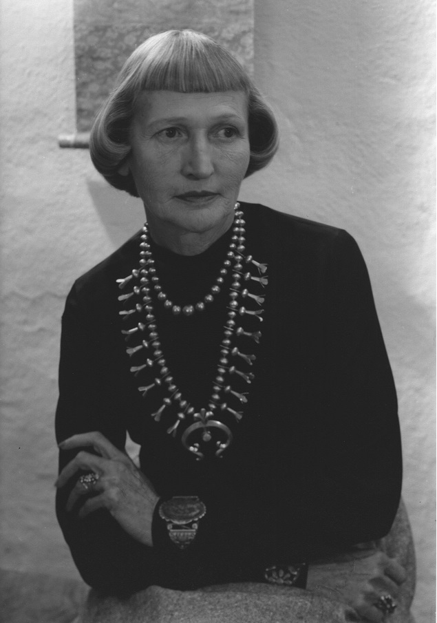 A black and white photo of a woman sitting in front of a textured white wall. She has a short bob haircut and straight bangs and is wearing a dark long-sleeved top and a large statement necklace. She has her arms crossed in front of her, resting on her lap. She wears a large cuff bracelet on each wrist and ornate rings on each hand. She is looking to the side with a neutral expression.