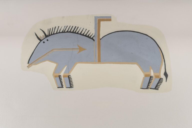 A photograph of a stylized, abstract painting of a blue animal with two humps and four legs on a neutral background. The animal is pictured in profile, but its two small black dots for eyes are both pictured on the side of its head, as are two small tan ears and two curved black horns. All four legs are also pictured. The top of the figure is outlined in black and it has a pointy black main and thin black tail. The bottom of the figure is outlined in tan. A thin brown and tan bent arrow points from its mouth to its stomach. An upside down L-shape made up of blue and brown lines extends upwards from its stomach and its back, rising in between the two humps.