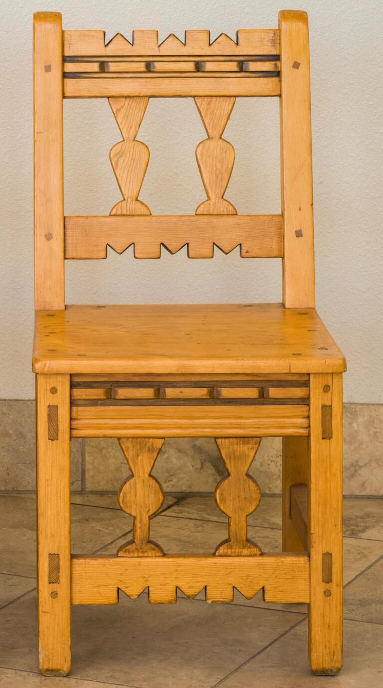 A photograph of a honey-colored wooden chair seen from straight on with almost identical arrangements of carved geometric designs on the backrest and base. The chair's design features gouged rectangular forms and inserts, M-shaped cut-outs and slats with vase-like shapes. The chair does not have armrests and is placed on a beige-tiled floor against a plain white wall.