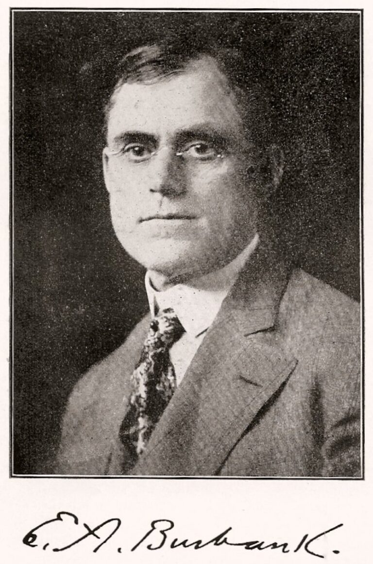 A black and white photograph of a man in a suit with a patterned tie and glasses. He is seen from the chest up, with short hair and a neutral expression. He looks in the direction of the camera with his gaze off in the distance. The image includes a handwritten signature at the bottom.