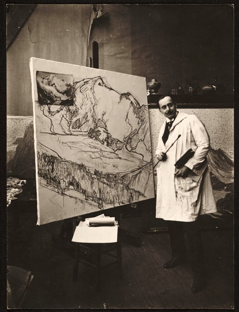 A sepia-toned photograph of a man with short dark hair and a mustache in a long white coat standing beside a large canvas with sketches of a mountain range. The man holds a paint brush in his right hand and leans slightly towards the canvas while looking away from it. Two paintings are partially visible propped up on the floor behind the man. We glimpse a table with paint palettes off to the side. And between the man and the canvas is a stool stacked with a disheveled pile of white papers. The room has high ceilings and an air of spaciousness.
