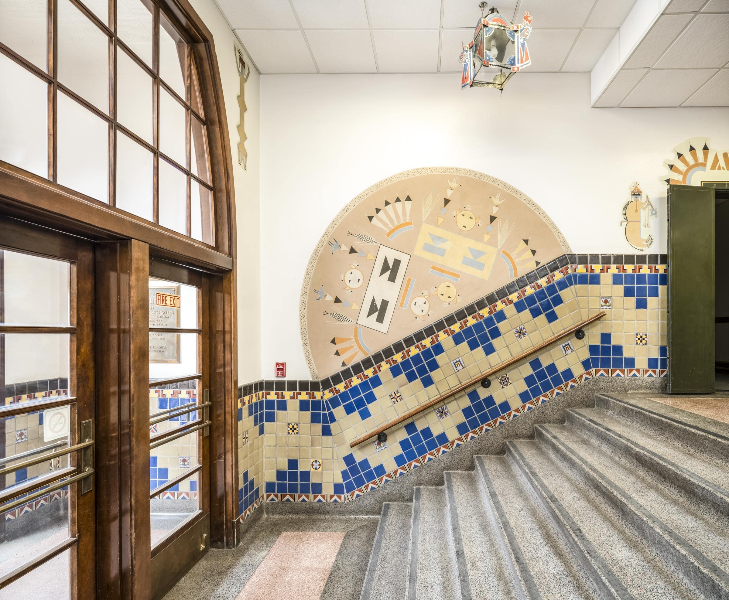 A photograph of a semicircular wall mural done in the style and using the visual language of Navajo sandpainting. The mural is positioned on a plain white wall at an angle, its bottom edge aligning with the top edge of the wall's tile wainscoting, which itself is angled to rise with the 8-step staircase in the foreground. The background of the mural is tan and it features a symmetrical design of geometric shapes. On the right half of the mural, a large light yellow rectangle is oriented 45 degrees to the left. The rectangle is decorated with two identical sets of shapes: a light blue triangle, point down, on top of a light blue rectangle. Above and below the rectangle are two yellow circular faces, with small black triangles for eyes and small black rectangles for mouths. On either side of the upper face are two stacks of narrow, downward-pointing triangles with stylized songbirds perched on top. And on either side of the triangle stacks are stylized ears of yellow corn. Mirroring this arrangement of shapes on the right side of the semicircle is the same pattern, angled 45-degrees in the opposite direction and done in black and white instead of blue and yellow. Stylized sets of three or five straight, angular feathers in yellow, white and orange are interspersed between these primary patterns.