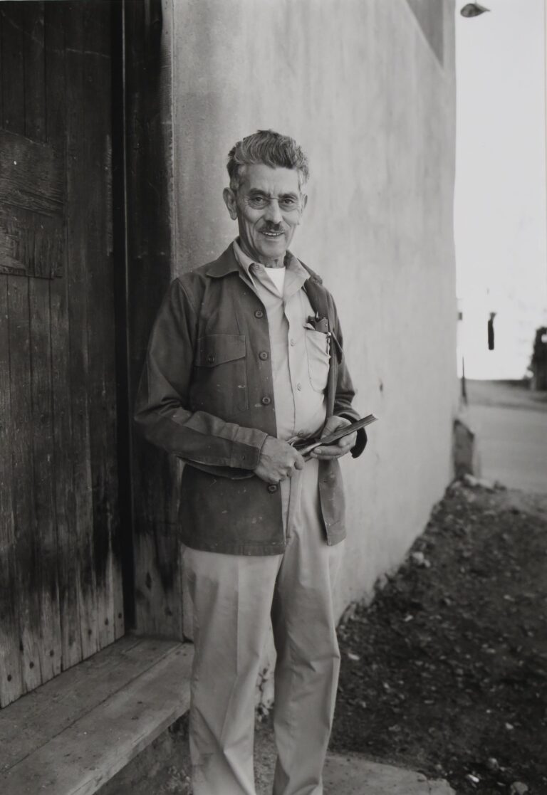 A black and white photograph of an older man with gray hair and a mustache standing outside of a building. He is wearing a jacket over a collared shirt, holding a chisel in his hands. He has a pocket protector or glasses case in his shirt pocket. The building behind him has a large wooden door with a wooden step.