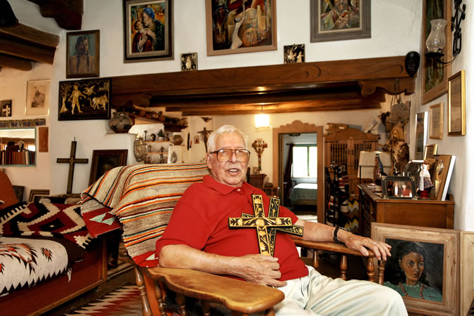 A color photo of an older man wearing a red shirt and beige pants sitting in a wooden rocking chair holding two crosses decorated with straw appliqué against his chest. He smiles at the camera wearing orange-tinted large square classes. The room is filled from floor to ceiling with artwork and decorations, including weavings, paintings, wood furniture, and other items.