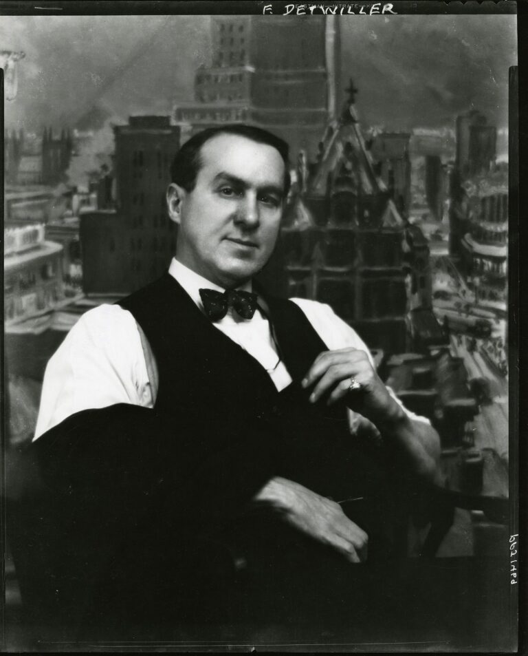 A black and white photo of a man with combed, short dark hair wearing a white shirt, bow tie, and a dark vest. The man looks confidently at the camera. He poses in front of a painted with a cityscape backdrop featuring tall buildings.