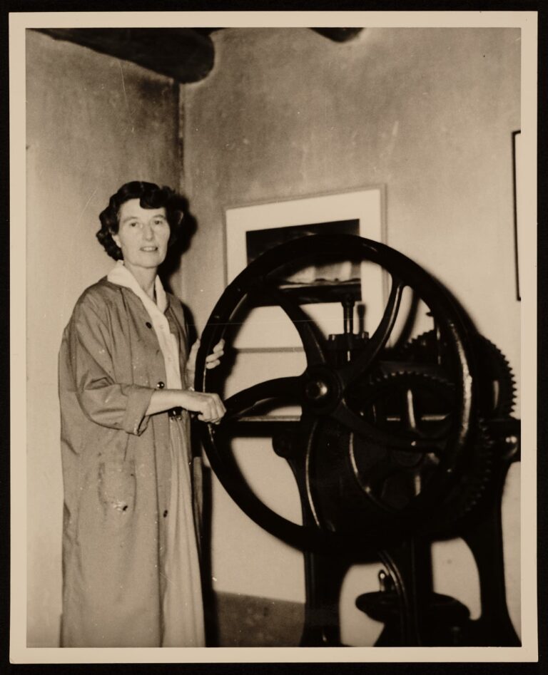 A black and white photograph of a woman with short brown hair wearing a long coat and standing beside a large mechanical wheel with visible gears. The machine is almost as tall as the woman and she has one hand holding its crank handle and the other gripping the rim of the wheel. She stands in a room, a framed artwork on the wall behind her and the ceiling's wood beams barely visible high above her.