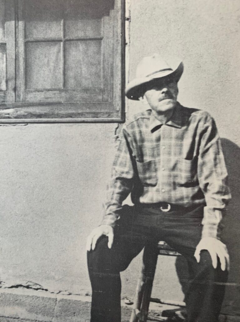 A black and white photograph of man in a well-worn cowboy hat and checkered collared shirt sitting on a stool outside a building with a wood-framed window over his right shoulder. He sits with his legs apart, a hand resting on each knee. He gazes up and to his left.