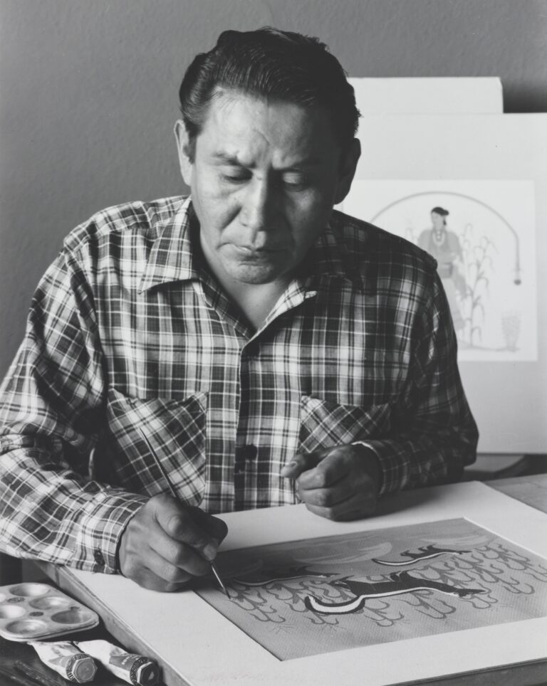 A black and white photo of a man with slicked back black hair wearing a plaid shirt and sitting at a table painting. He appears focused, holding a thin paintbrush directly above his paper, on which a scene of skunks and cornstalks is painted. A palette and two paint tubes sit on the table next to him, and a painting of a woman in a field of corn is partially visible behind his left shoulder.