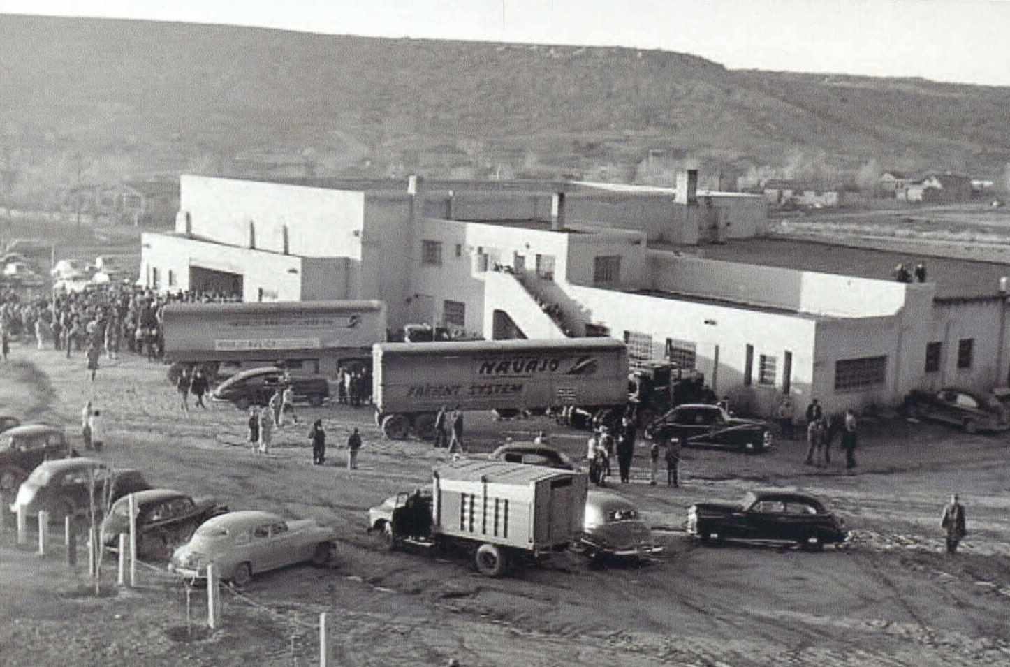 A vintage black-and-white photo shows a large building with two trucks labeled Navajo Freight Lines parked in front. Numerous people and cars are gathered around the building, with a hilly landscape in the background.