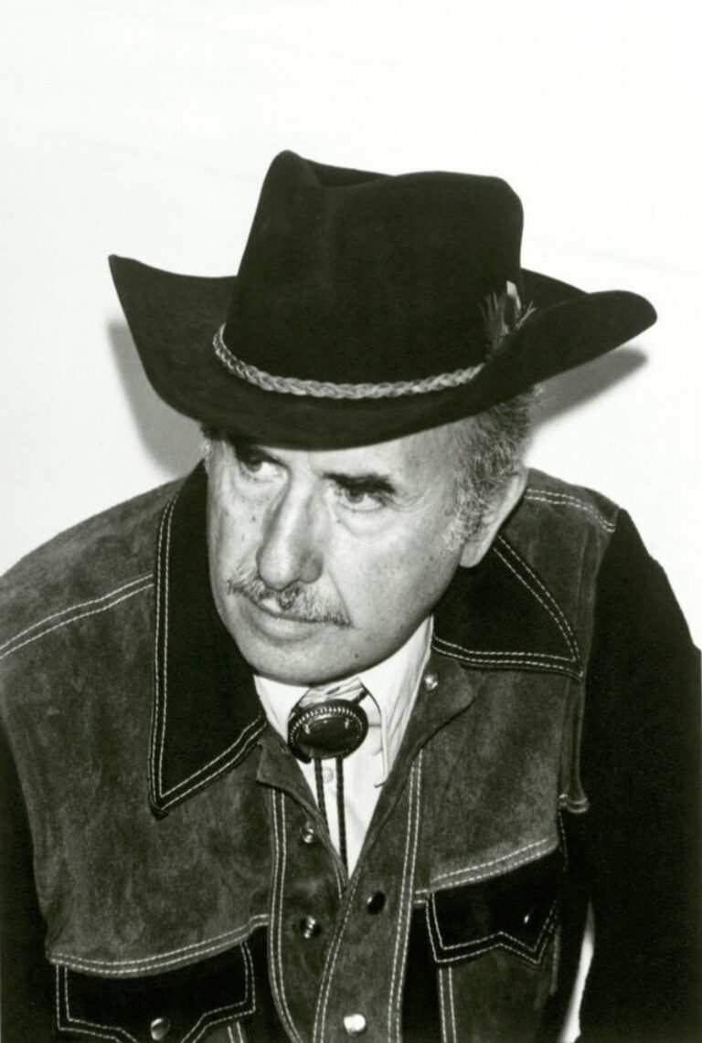 A black and white photograph of an older man with a mustache wearing a black cowboy hat with a decorative band, western-style jacket with visible white stitching, and a bolo tie. The photograph appears take from slightly above, with the man posed leaning forward and looking off to the side.
