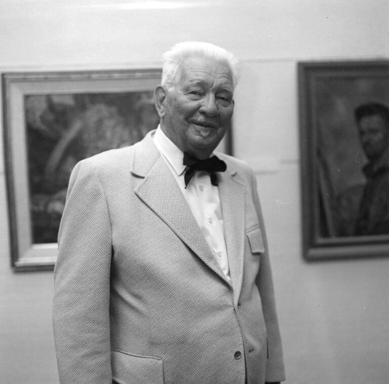 A black and white photograph of older man with white hair, wearing a light-colored suit and a black bow tie, standing in a gallery setting. He is seen from the waist up and is looking at the camera and smiling, and there are framed paintings on the wall behind him.