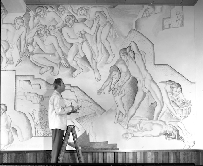 A black and white photograph of an artist standing on a ladder painting a large mural. The mural depicts various human figures in dynamic poses. The artist is seen in profile, looking at the mural, holding multiple paint brushes in his hands, as if contemplating his next move. He wears a waist-length white coat and slacks. The artist appears slightly smaller than the human figures in his painting.