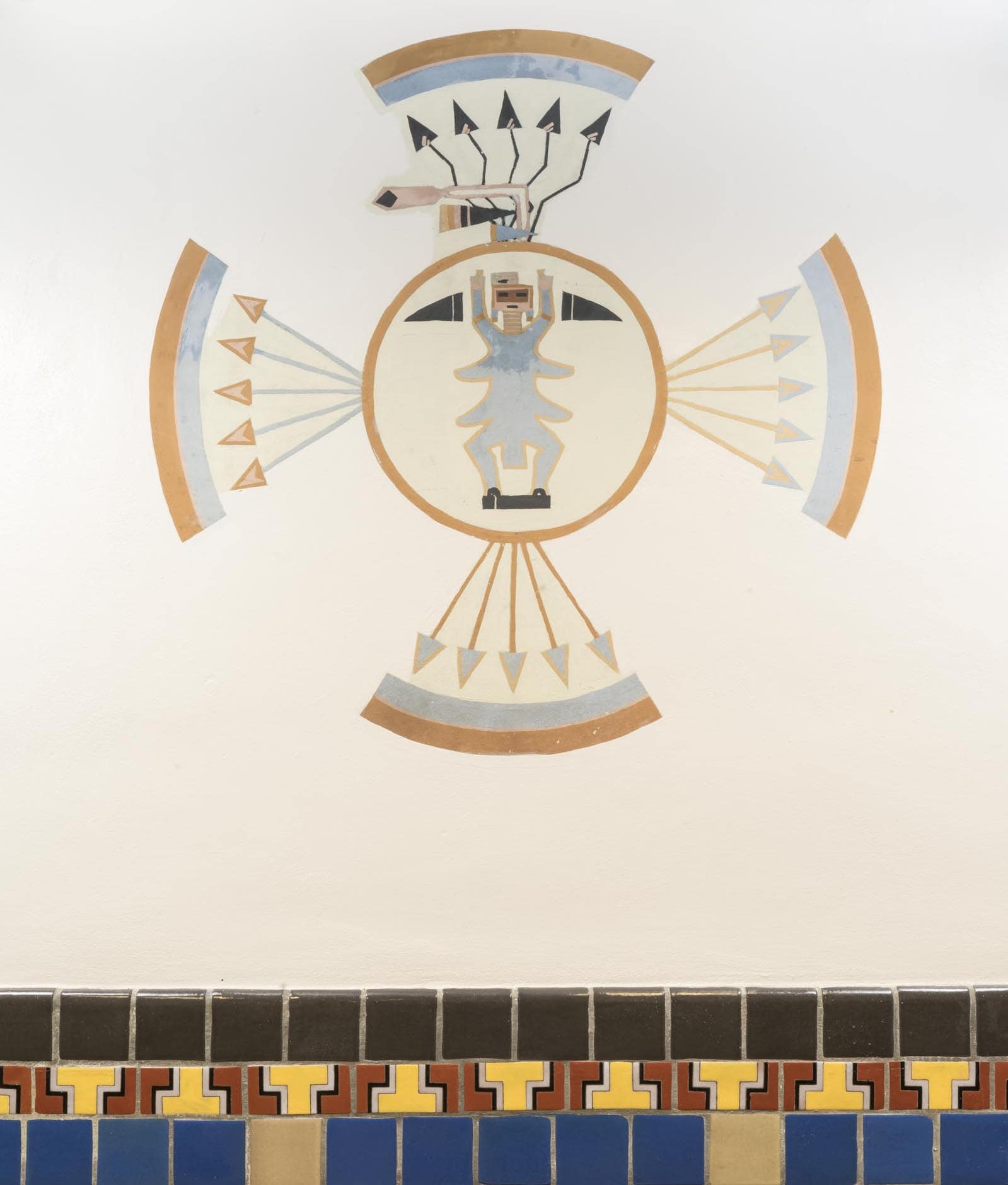 A photograph of a wall painting done in the style and using the visual language of Navajo sandpainting, done on a plain white wall above decorative tile wainscoting. The wall painting design is highly symbolic and centers on a yellow circle outlined in orange with a stylized blue figure in the middle. The figure has a rectangular head, small black rectangles for the eyes and mouth and two flap-like protrusions on either side of its torso. Four sets of five arrows protrude from the circle to the left, right, up and down, aligning with the cardinal directions. The arrows start close together and fan out, pointing to a curved band of color, half blue and half orange. The arrows all have straight staffs, except for the top ones, which are black and have zig-zagged staffs.