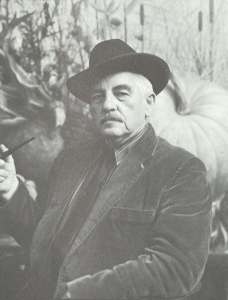 A black and white photograph of an older man with a thick mustache and dark fedora-style hat. Seen from the waist up, the man appears to be sitting at a slight angle, resting his front arm on the frame of his chair and holding a pipe with his other hand. He turns his head to look sternly at the camera. He wears a striped collared shirt and velvet jacket and poses against what appears to be a background painted with a large pumpkin, gourd and tall grasses.