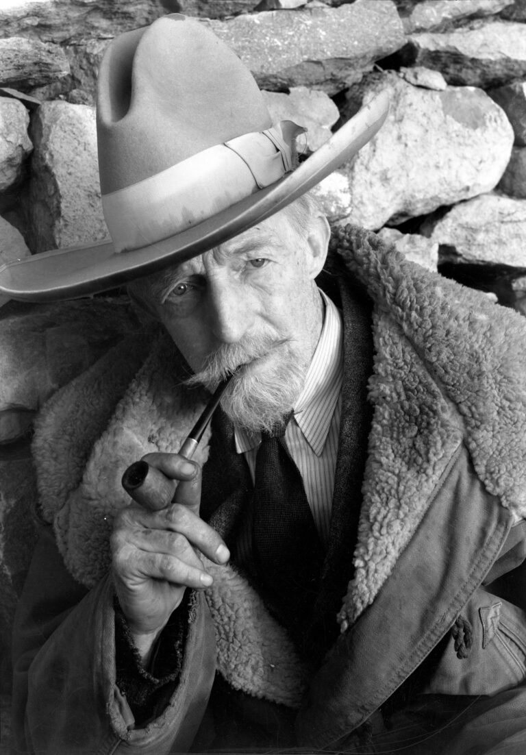 A black and white photograph of an older man with a thick mustache and goatee holding a pipe between his lips. He is seen at close range, posing in front of a rock wall and wearing a tall, wide-brimmed cowboy hat with satin trim, a shirt and tie, and a sheepskin-lined coat. He gazes directly at the camera with a thoughtful expression.