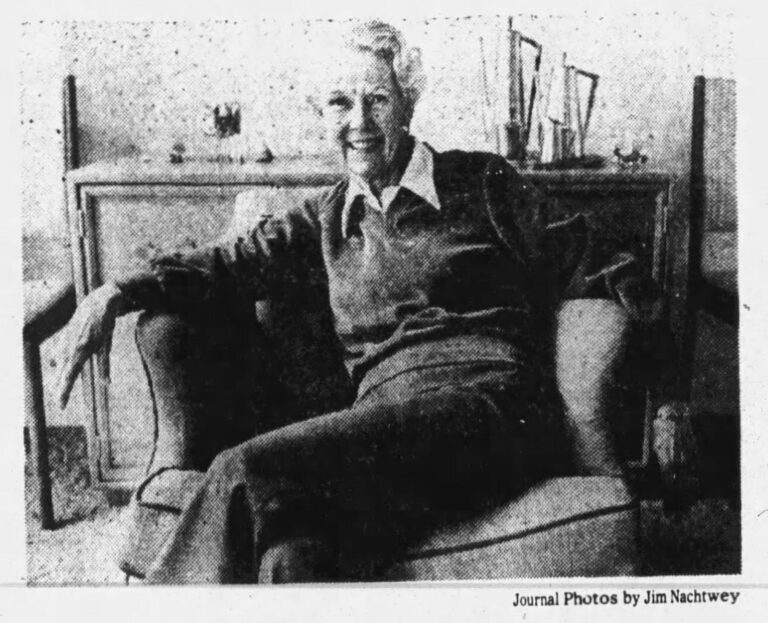 A black and white photograph of a woman with short white hair sitting casually on an upholstered armchair, wearing a sweater over a collared shirt, and smiling at the camera. Her demeanor is relaxed as she crosses her legs, leans back, and lets her arms fall over the chair's armrests. There is a cabinet in the background with some decorative items.