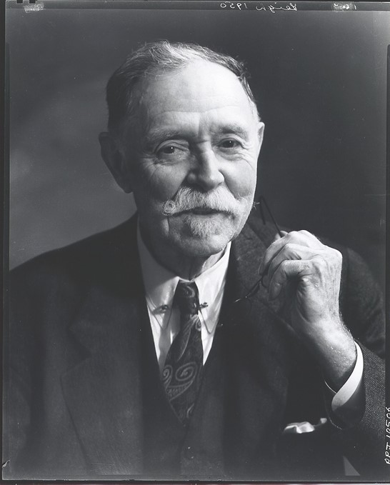 A black and white portrait of a smiling older man with a curled white mustache. Seen from the chest up, he wears a three-piece suit with a tie and holds his glasses in one hand. The lighting of the photograph highlights the man's hand and face, and his gaze is warm and friendly.