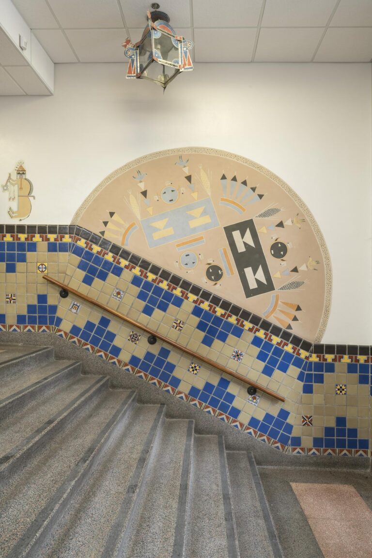 A photograph of a semicircular wall mural done in the style and using the visual language of Navajo sandpainting. The mural is positioned on a plain white wall at an angle, its bottom edge aligning with the top edge of the wall's tile wainscoting, which itself is angled to rise with the 8-step staircase in the foreground. The background of the mural is tan and it features a symmetrical design of geometric shapes. On the left half of the mural, a large light blue rectangle is oriented 45 degrees to the left. The rectangle is decorated with two identical sets of shapes: a light yellow triangle, point down, on top of a light yellow rectangle. Above and below the rectangle are two light blue circular faces, with small black triangles for eyes and a small black rectangles for mouths. On either side of the upper face are two stacks of narrow, downward-pointing triangles with stylized songbirds perched on top. And on either side of the triangle stacks are stylized ears of yellow corn. Mirroring this arrangement of shapes on the right side of the semicircle is the same pattern, angled 45-degrees in the opposite direction and done in black and white instead of blue and yellow. Stylized sets of three or five straight, angular feathers in yellow, blue and orange are interspersed between these primary patterns.