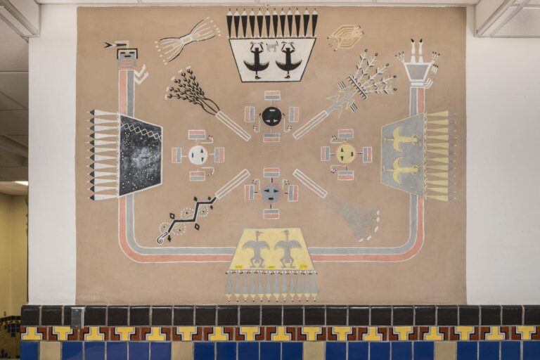 A photograph of an intricate mural on a wall done in the style and using the visual language of a Navajo sandpainting. The painting is an intentionally ordered combination of specific and detailed symbols on a rectangular tan background. The overall design is oriented to the four cardinal directions. The perimeter of the design features four trapezoidal shapes, one in the center of each side, and each painted in one of the four sacred Navajo colors: white, black, blue and yellow. All of the trapezoids have a neat row of straight feathers extending out from their bases. The white, blue and yellow trapezoids contain stylized bird figures and the black trapezoid is painted with white dots to represent stars or constellations. The trapezoids are connected on the left, right, and bottom sides by the elongated U-shaped torso--made up of one red band and one blue band--of a figure whose rectangular head is positioned in the upper left corner of the image and whose skirt and legs are positioned upside down in the upper right. The interior of the painting is made up of four circular faces, each with small black triangles for eyes and a small black rectangles for mouths, aligned with the trapezoids and also painted one of each sacred color. In between the circular faces, stylized depictions of the four sacred Navajo plants, tobacco, corn, beans, and squash, are oriented on a diagonal.