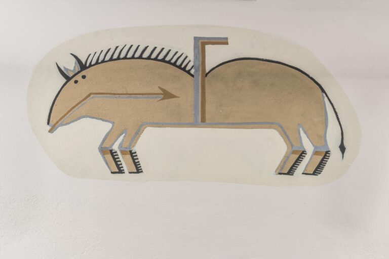 A photograph of a stylized, abstract painting of a tan animal with two humps and four legs on a neutral background. The animal is pictured in profile, but its two small black dots for eyes are both pictured on the side of its head, as are two small tan ears and two curved black horns. All four legs are also pictured. The top of the figure is outlined in black and it has a pointy black main and thin black tail. The bottom of the figure is outlined in blue. A thin brown and blue bent arrow points from its mouth to its stomach. An upside down L-shape made up of blue and brown lines extends upwards from its stomach and its back, rising in between the two humps.
