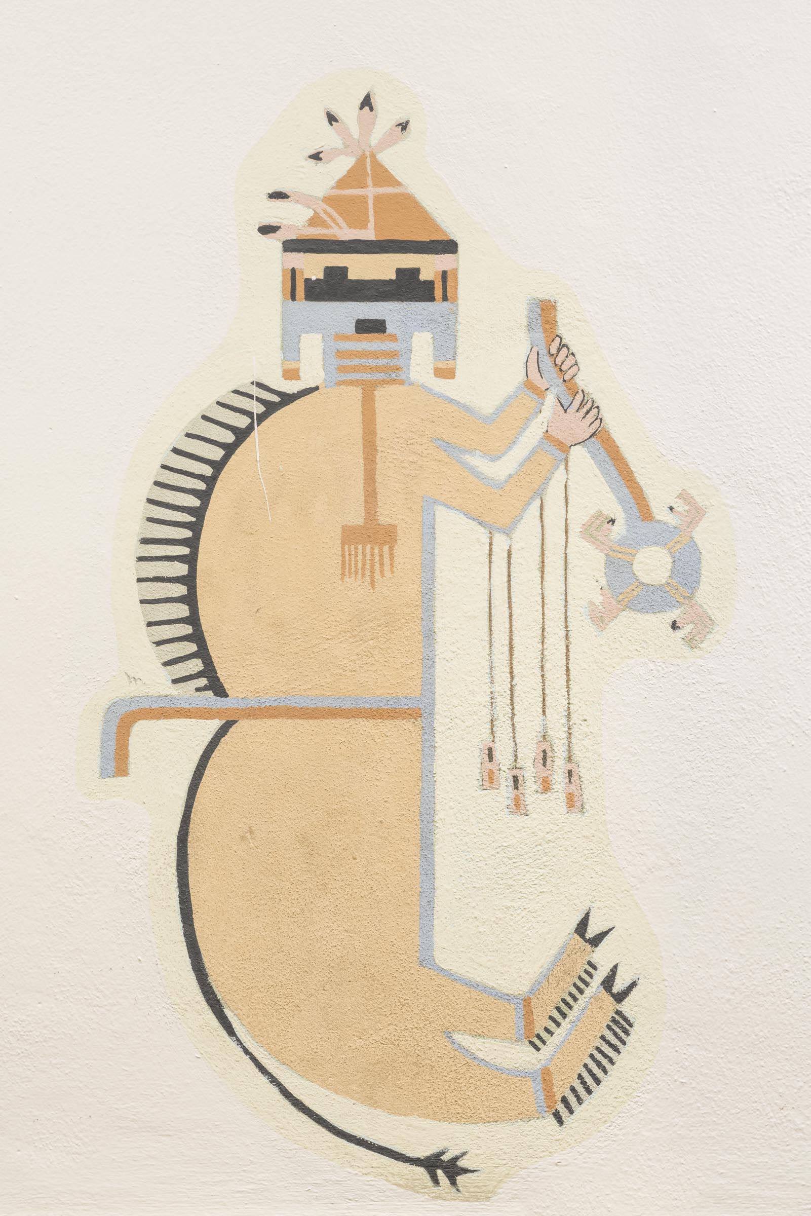 A photograph of a painting of a stylized, tan-colored figure on a light background. The figure is oriented vertically. It has a rectangular head with small black rectangles for the eyes and mouth and wears a light brown triangular hat adorned with feathers. It has two humps on its back, a pointy black mane, a thin black tail, and hoofed feet. Its arms are bent in front of its body and two peach-colored human hands hold a staff with a circular piece at its bottom.