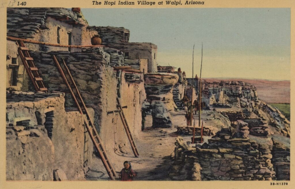 A vintage postcard showing the Hopi Indian Village at Walpi, Arizona, as the text at the top reads. The image is a colored black and white photographic print and features stone dwellings in tan and gray shades built into the side of a cliff, with brown wooden ladders leading to different levels. Figures colored brown are depicted along a path near base of the structure. The background is a clear blue sky which fades to white along the horizon above a pinkish streak perhaps representing mountains or mesas, a small patch of green beneath it.