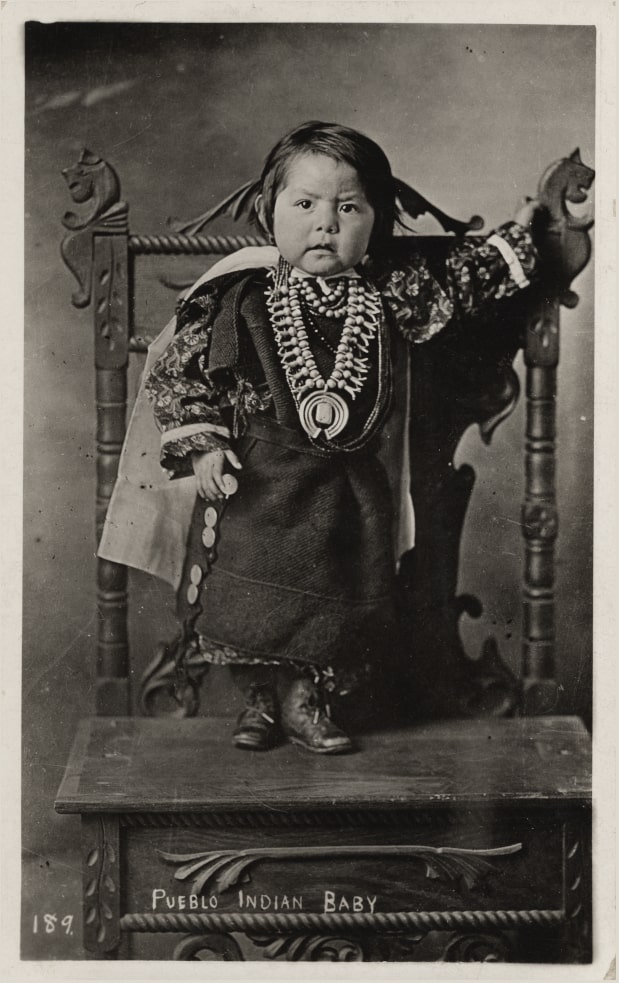 A historical black and white photograph of a toddler standing on an ornately carved large, wooden high-backed chair. The toddler is about as tall as chair's backrest and has short black hair and chubby cheeks. She is dressed in traditional regalia, including a black sleeveless dress with round silver side buttons worn over a long-sleeved floral patterned shirt. She also wears a white cape and a large silver necklace with a circular pendant. The child rests one hand on the back of the chair and holds the other by her side. Handwritten text at the bottom of the image reads "Pueblo Indian Baby."