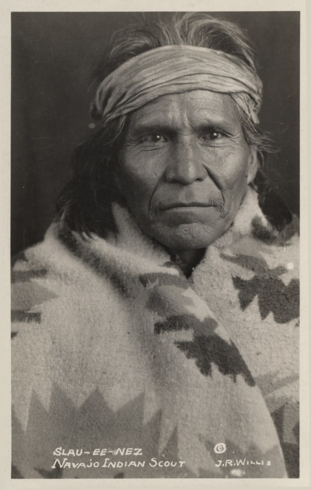 A historical black-and-white portrait of a man pictured from the chest up facing the camera with a distant look in his eyes and a solemn expression. He wears a patterned blanket wrapped around his shoulders and his shoulder-length white hair is wrapped in a headband tied around his forehead. Handwritten text on the bottom of the image reads "Slau-ee-nez Navajo Indian Scout." The photograph is signed "J.R. Willis" with a copyright symbol.
