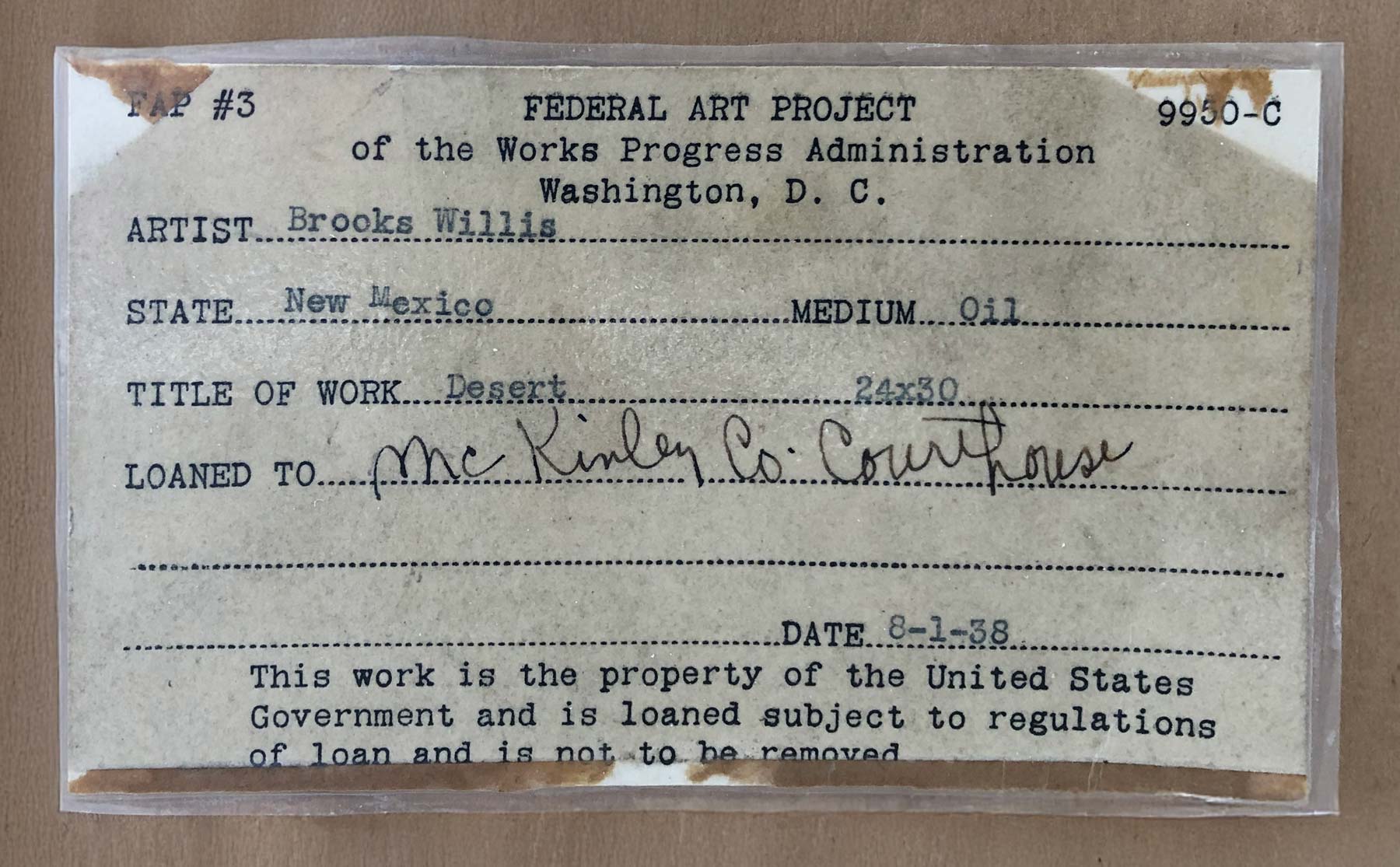 A laminated typewritten label from the Federal Art Project listing information about an artwork and a disclaimer that "the work is the property of the United States Government and is loaned subject to regulations of loan and is not to be removed." It includes spaces for the artist's name (filled in with Brooks Willis), the state (New Mexico), the medium (oil), the title of work (Desert), and a loan note to "McKinley Co. Courthouse." The label also includes the dimensions (24 x 30) a date of 8-1-38.