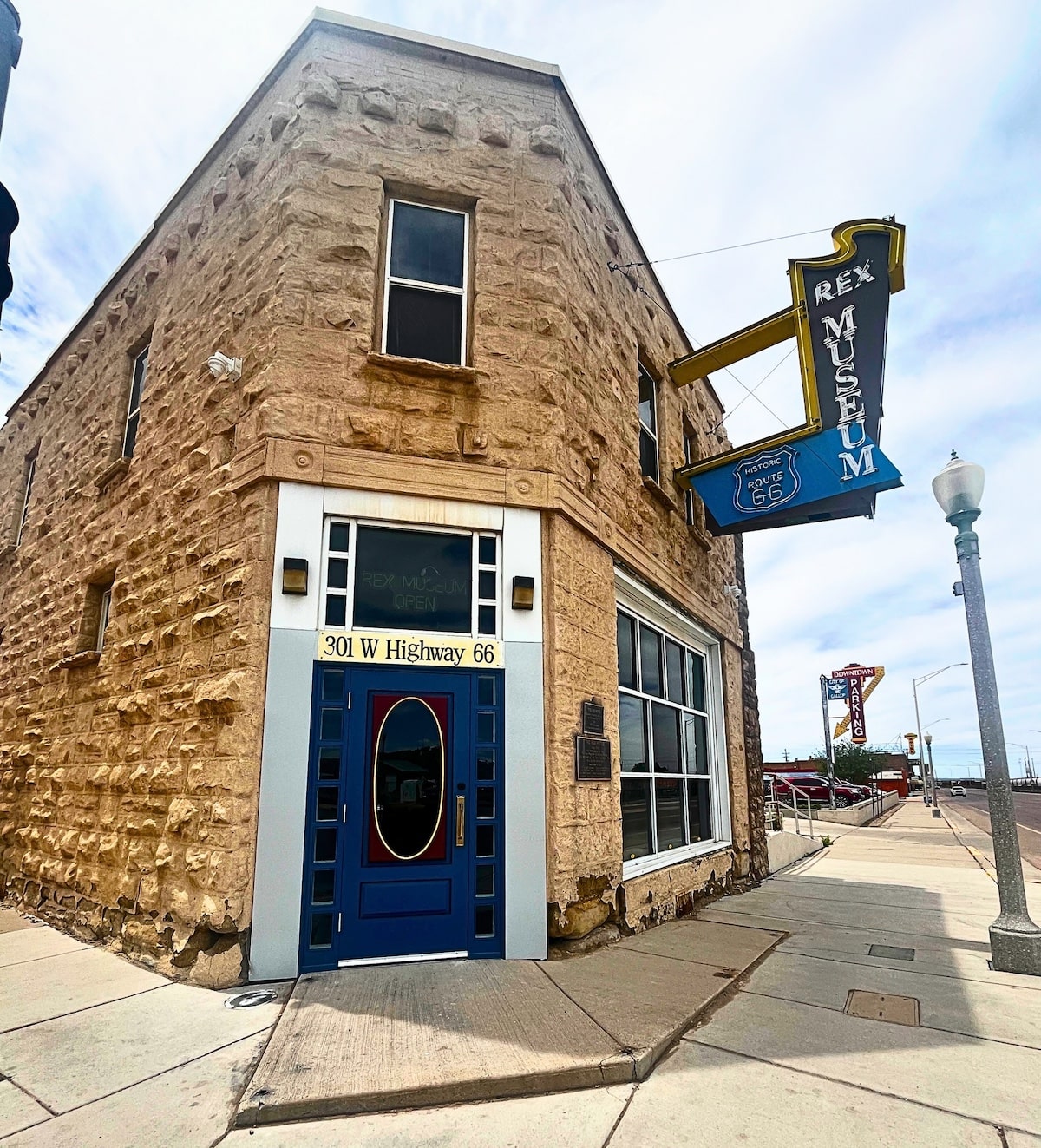 A photograph of the exterior of the Rex Museum, the sign for which projects off the wall to the left of the main entrance. It is done in a vintage neon design. The two-story building is built from rough sandstone blocks and is oriented so the doorway is angled to the street corner. The door is blue with an oval window and is lined by paned glass. There is a large street-level window to the right of the door and small rectangular windows line the second level.