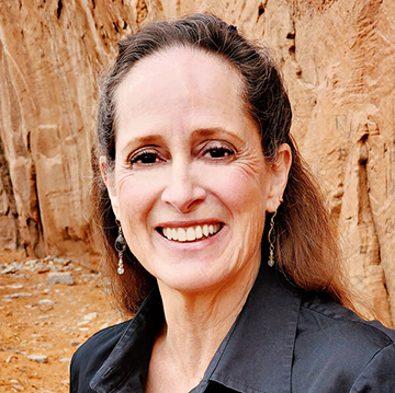 A photograph of a woman, pictured from the shoulders up, wearing a black collared shirt, with long reddish-brown hair, brown eyes, and dangling earrings. She is smiling at the camera and standing in front of a tan-colored cliff or natural rock wall.